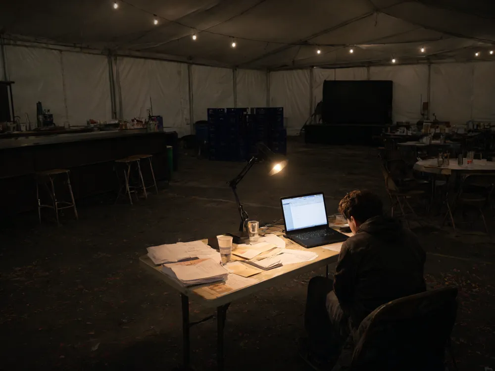 A volunteer sits alone at night in an empty festival tent at a table full of timesheets